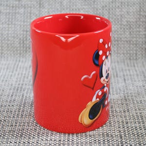 Peut inclure: Mug rouge en c&eacute;ramique avec Minnie Mouse. Le mug a une finition brillante et une forme cylindrique. Minnie Mouse est repr&eacute;sent&eacute;e dans sa tenue classique avec un n&oelig;ud &agrave; pois et des gants. Un c&oelig;ur est &eacute;galement en relief sur le mug.