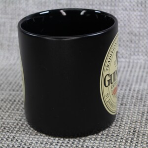 May include: A black ceramic mug with a cylindrical shape. The mug features a cream-colored label with the Guinness logo and text. The mug is designed for beverages and is suitable for home or bar use.