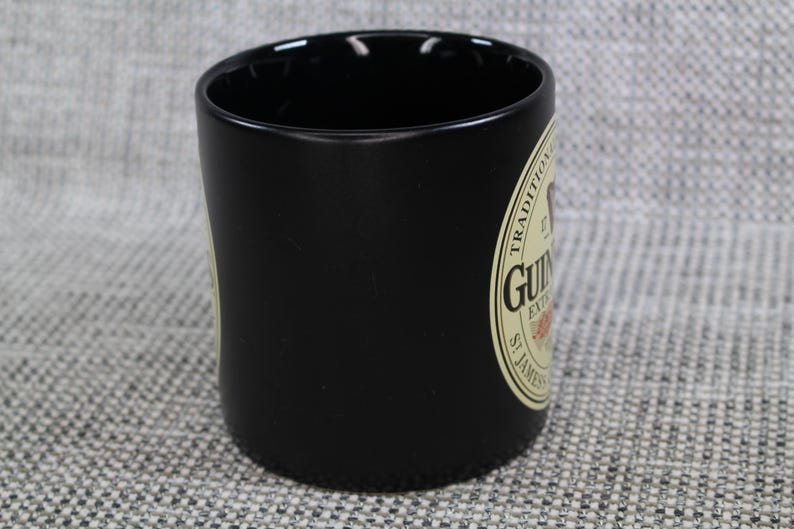 May include: A black ceramic mug with a cylindrical shape. The mug features a cream-colored label with the Guinness logo and text. The mug is designed for beverages and is suitable for home or bar use.