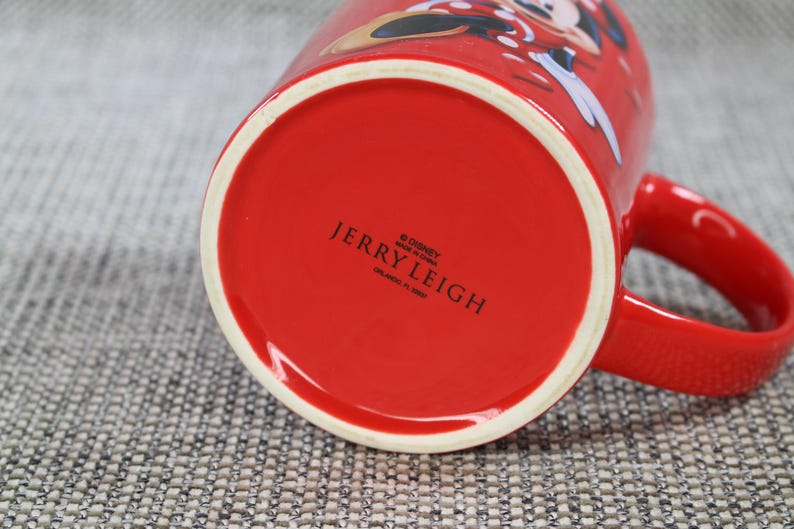 Peut inclure: Une tasse en c&eacute;ramique rouge avec un bord blanc et une anse rouge. Le fond de la tasse porte l'inscription "JERRY LEIGH" et le logo Disney. La tasse pr&eacute;sente un motif de dessin anim&eacute; sur le c&ocirc;t&eacute;.