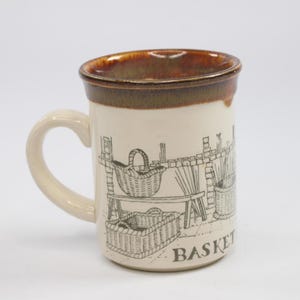 May include: A ceramic mug with a cream-colored body and a brown rim. The mug features a black and white illustration of baskets and the word "BASKET" on the side. The handle is cream-colored.
