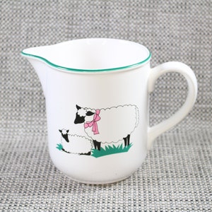 May include: White ceramic pitcher with a green rim and a black and white sheep design. The sheep has a pink bow. The pitcher has a handle.