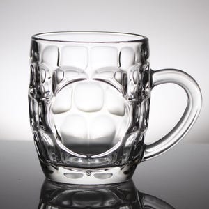 May include: A clear glass beer mug with a handle. The body of the mug has a textured, dimpled pattern. The handle is a curved loop. The mug is empty and sits on a reflective surface.
