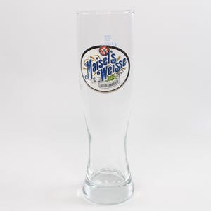Puede incluir: Un vaso de cerveza alto y transparente con forma curva. El vaso presenta un logotipo ovalado blanco y azul con las palabras "Maisel's Weisse" y "AUS BAYREUTH". El vaso está vacío y sobre un fondo blanco.