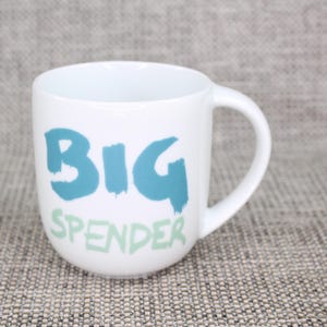 May include: A white ceramic mug featuring the text BIG SPENDER printed on the front. The word BIG is in a large, bold, teal brushstroke font, while the word SPENDER is in a smaller, lighter green brushstroke font below it.