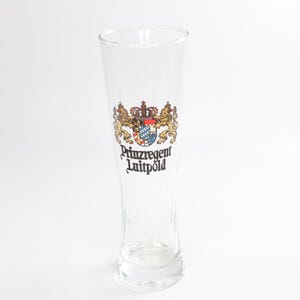 Puede incluir: Un vaso de cerveza transparente y alto con la parte superior ensanchada. El vaso presenta un emblema colorido con una corona, un escudo y el texto "Prinzregent Luitpold". El vaso está diseñado para cerveza y es adecuado para servir.