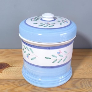 May include: A blue and white ceramic canister with a lid. The canister features a floral design with green leaves and pink flowers. The lid has a white knob.