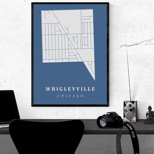 May include: A framed map print of Wrigleyville, Chicago, in a minimalist style. The map is white on a blue background, with the neighborhood name and city below. The print is displayed on a desk with a laptop, camera, and other items.