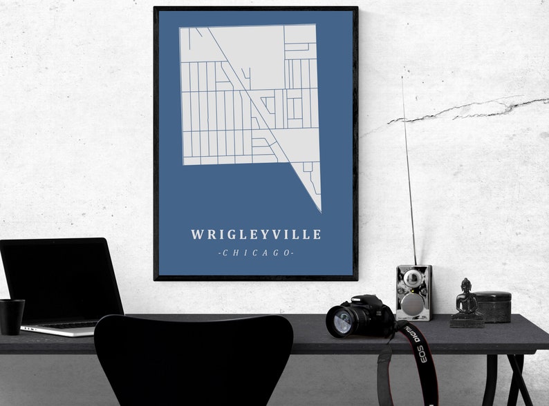 May include: A framed map print of Wrigleyville, Chicago, in a minimalist style. The map is white on a blue background, with the neighborhood name and city below. The print is displayed on a desk with a laptop, camera, and other items.