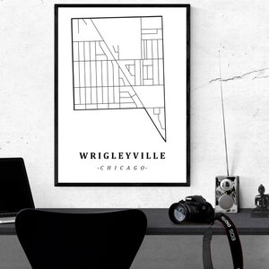 May include: A black and white framed print of a map of Wrigleyville, Chicago. The print is displayed on a white wall above a desk with a laptop, camera, and other items. The text "Wrigleyville - Chicago" is at the bottom.