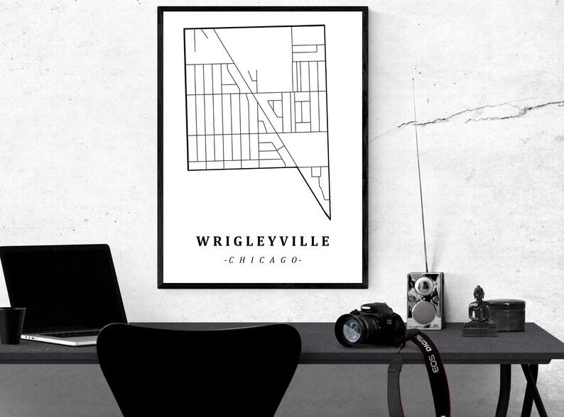 May include: A black and white framed print of a map of Wrigleyville, Chicago. The print is displayed on a white wall above a desk with a laptop, camera, and other items. The text "Wrigleyville - Chicago" is at the bottom.