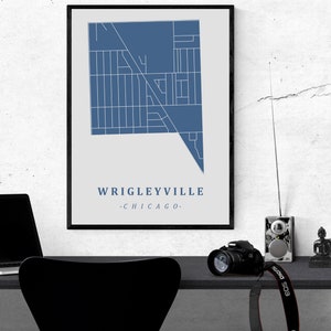 May include: A framed print featuring a blue and white map of Wrigleyville, Chicago. The text "Wrigleyville - Chicago" is displayed below the map. The print is displayed on a desk with a laptop, camera, and other items.