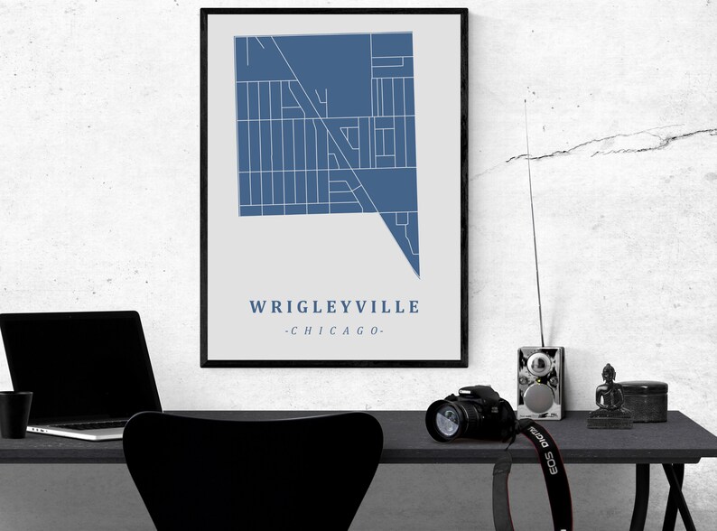 May include: A framed print featuring a blue and white map of Wrigleyville, Chicago. The text "Wrigleyville - Chicago" is displayed below the map. The print is displayed on a desk with a laptop, camera, and other items.
