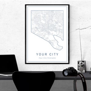 May include: A framed map print of a city, with a blue outline and details on a white background. The text "YOUR CITY" is below the map. A laptop, camera, and other items are on a black desk.