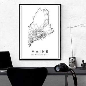 May include: A framed black and white print of the Maine state map, showing road networks. The word "MAINE" is below the map, with "-The Pine Tree State-" underneath. The print hangs on a white wall, with a desk and accessories in the foreground.