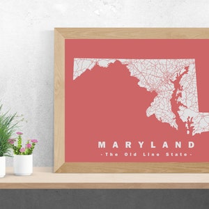 May include: A framed print featuring a map of Maryland in white lines against a coral background. The word "MARYLAND" is displayed in white text, with "- The Old Line State -" below it. The frame is made of light-colored wood.