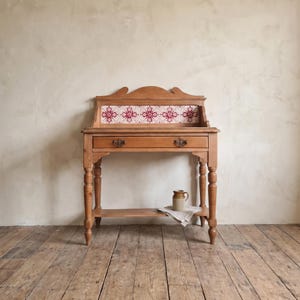 May include: A light brown wooden console table featuring a single drawer with two dark metal pulls. The back panel is decorated with a row of white and red patterned tiles. A small beige ceramic pitcher sits on the lower shelf atop a folded cloth.