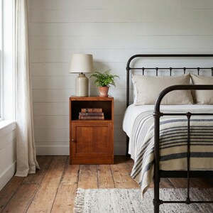 May include: A bedroom with a black metal bed frame, striped blanket, and pillows. A wooden bedside cabinet holds a lamp, books, and a plant. The room has wooden flooring and a white, slatted wall. The lamp is beige.