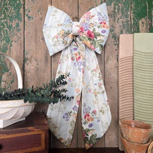 May include: A large decorative bow made of floral fabric, with a light blue striped background and colorful flowers. The bow is attached to a gold ring and hangs against a rustic wooden background. The bow's design includes pink, yellow, and purple flowers.