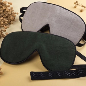 Puede incluir: Dos máscaras para dormir, una gris y otra verde oscuro, con correas elásticas negras. Las correas tienen un patrón ondulado.