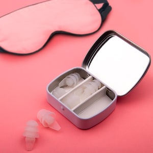 May include: A white travel case with a mirror lid, containing multiple pairs of clear earplugs. The case is open and sitting on a pink surface. There is a pink sleep mask in the background.