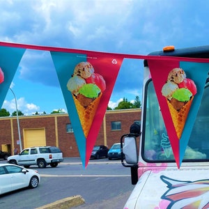 May include: A pink ice cream truck with a banner of ice cream cones hanging above it. The banner is made of red, blue, and white triangular flags with images of ice cream cones on them. The truck is parked in front of a brick building.
