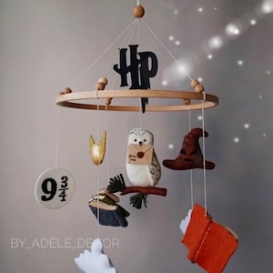 May include: A handmade baby mobile featuring felt Harry Potter-themed elements. The mobile includes a wooden ring, a black "HP" logo, a golden snitch, an owl with a letter, a sorting hat, a book, a platform 9 3/4 sign, and a white ghost.