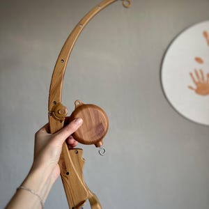 May include: Wooden mobile arm with a rotating wooden disc. The arm is brown and has a metal hook at the end. The disc is also brown and has a wood grain pattern.