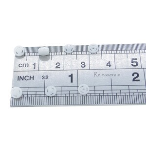 May include: White plastic buttons with a raised design, measured against a ruler in inches. The buttons are approximately 1/2 inch in diameter.