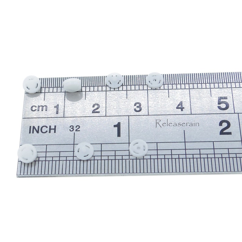 May include: White plastic buttons with a raised design, measured against a ruler in inches. The buttons are approximately 1/2 inch in diameter.
