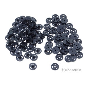 May include: A large pile of black plastic snap buttons, with a smaller pile of the same buttons to the right. The buttons are round with a raised center and three concentric circles.
