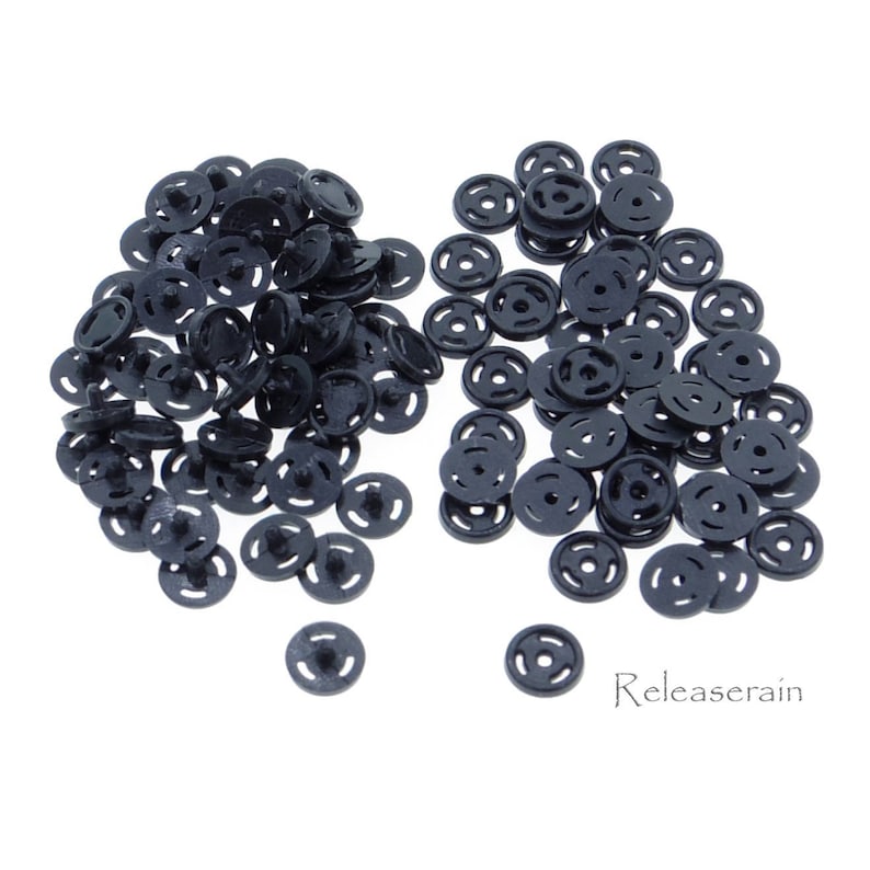 May include: A large pile of black plastic snap buttons, with a smaller pile of the same buttons to the right. The buttons are round with a raised center and three concentric circles.