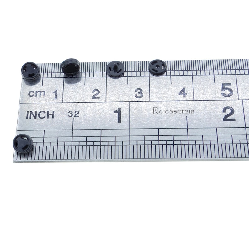 May include: Five black plastic buttons, each with a different design, are arranged on a ruler. The ruler is marked in centimeters and inches. The buttons are approximately 1/2 inch in diameter.