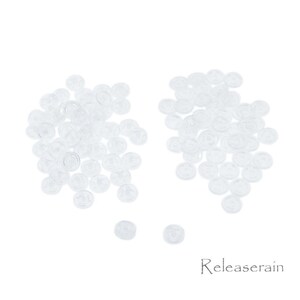 May include: A large pile of clear plastic buttons, with a smaller pile of buttons to the right. The buttons are all the same size and shape, and they are arranged in a random pattern.
