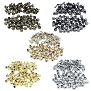 Könnte beinhalten: Sortiment aus Metallnieten in fünf verschiedenen Farben: Gold, Silber, Bronze, Schwarz und Nickel. Die Nieten sind in fünf Stapeln auf weißem Hintergrund angeordnet.