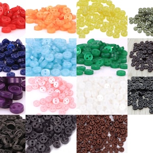 Pode incluir: Conjunto de 12 cores de botões de plástico de dois furos. Os botões são pequenos e redondos, com um acabamento liso. As cores incluem vermelho, laranja, amarelo, verde, azul, roxo, rosa, branco, preto, marrom, verde escuro e azul escuro.