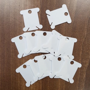 May include: A set of ten white plastic thread bobbins. The bobbins are designed to hold thread for embroidery, sewing, or other crafts.