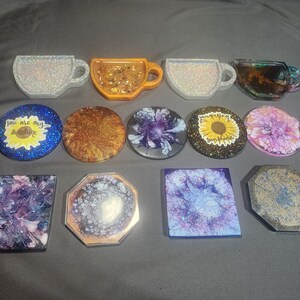 May include: A selection of resin coasters and cup-shaped items. The coasters are round, square, and octagonal, with glitter, swirls, and floral patterns. Some coasters have the text "You are my sunshine". The cup-shaped items are glittery.