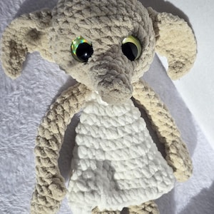 May include: A handmade crochet doll with large, iridescent eyes and oversized ears. The doll is primarily beige with a white dress. The arms and legs are also beige, and the doll has a long, pointed nose. The doll is made of a soft, textured yarn.