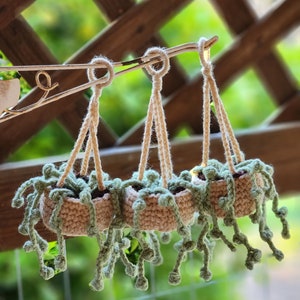 May include: Three crocheted hanging planters with green leaves and beige pots. The planters are hanging from a metal hook.