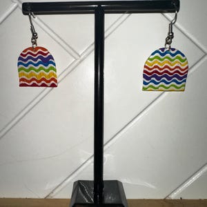 Könnte beinhalten: Ein Paar farbenfrohe Ohrringe mit einem Regenbogenwellenmuster. Die Ohrringe werden auf einem schwarzen Ohrringständer präsentiert. Die Ohrringe haben einen silbernen Haken und eine abgerundete Oberseite.