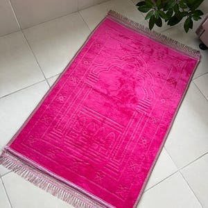 May include: A pink prayer rug with a decorative design and fringe. The rug has a traditional Islamic arch design in the center.