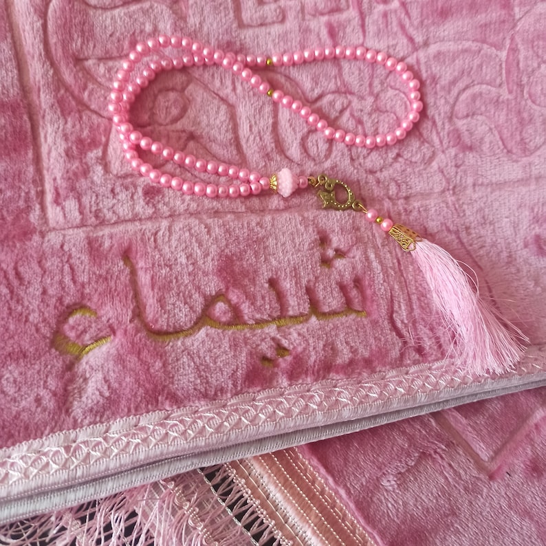 May include: A pink prayer rug with a gold embroidered Arabic inscription "Allah" and a pink prayer bead necklace with a gold tassel.