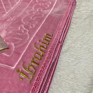 Pode incluir: Um tapete de ora&ccedil;&atilde;o rosa com bordados dourados que dizem "Ibrahim". O tapete tem uma borda rosa e branca.