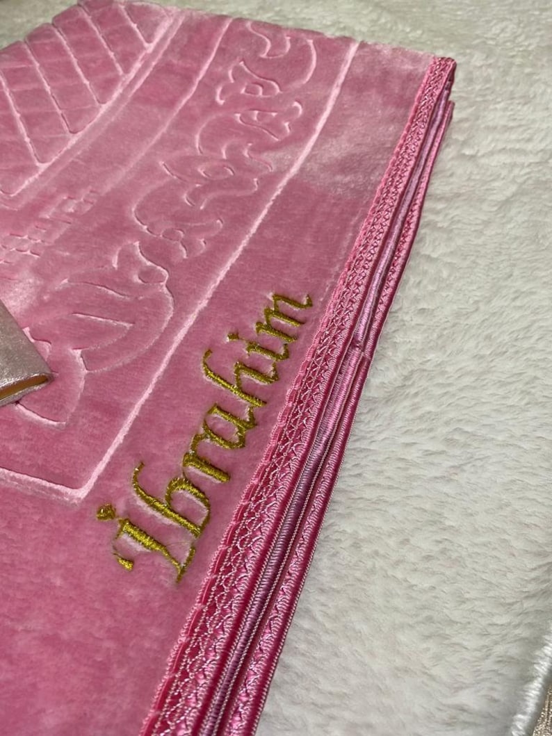 Pode incluir: Um tapete de ora&ccedil;&atilde;o rosa com bordados dourados que dizem "Ibrahim". O tapete tem uma borda rosa e branca.
