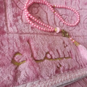 May include: A pink prayer rug with a gold embroidered Arabic inscription and a pink beaded prayer chain with a tassel.