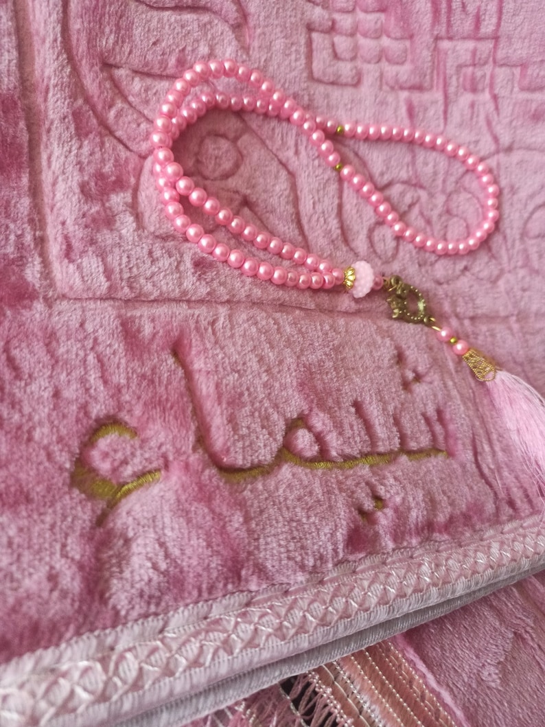 May include: A pink prayer rug with a gold embroidered Arabic inscription and a pink beaded prayer chain with a tassel.