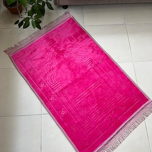 May include: A pink prayer rug with a decorative design and fringe. The rug is made of soft, plush fabric.