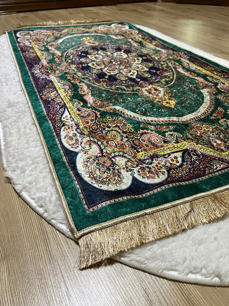 Peut inclure: Un tapis de pri&egrave;re vert avec une frange dor&eacute;e et des motifs floraux complexes. Le tapis est sur une surface blanche et moelleuse.