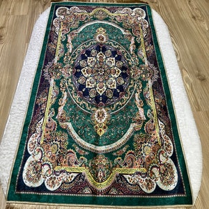Peut inclure: Un tapis de pri&egrave;re vert avec des motifs floraux et g&eacute;om&eacute;triques complexes dans des tons d'or, de jaune, de rouge et de bleu. Le tapis a une bordure de franges dor&eacute;es.